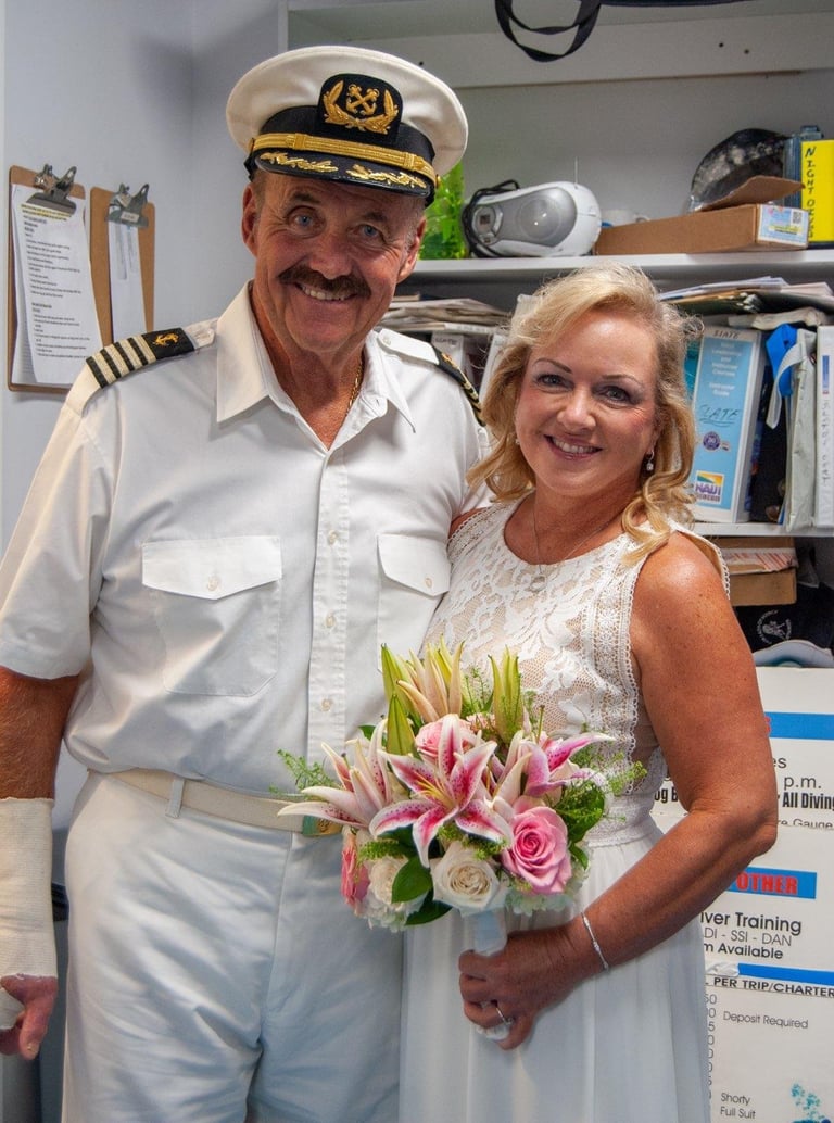 Man in captains uniform with mustache and woman holding a bouquet of pink lilies and roses posing together indoors