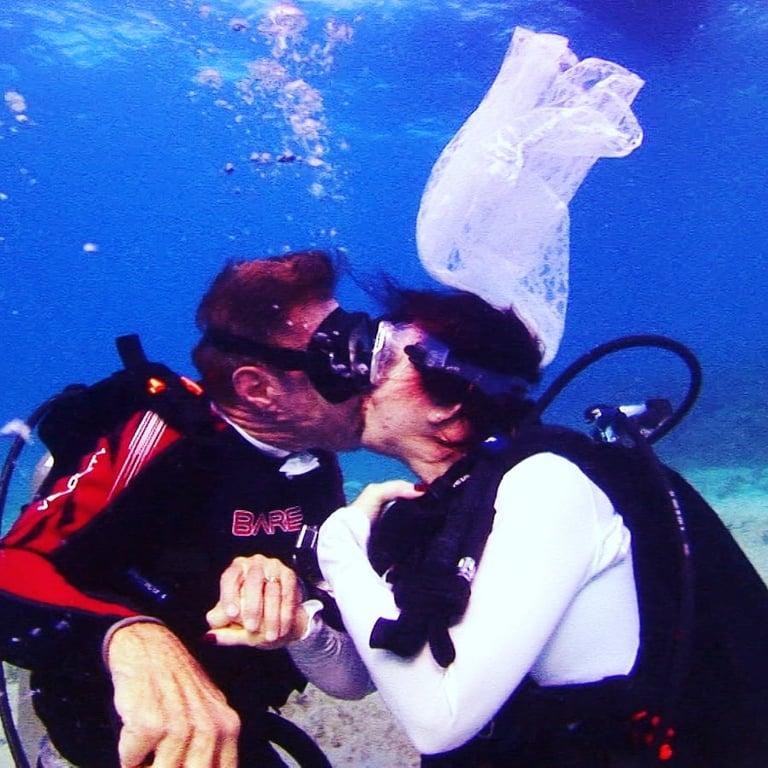 Two scuba divers underwater embrace while a plastic bag floats above them in clear blue ocean water