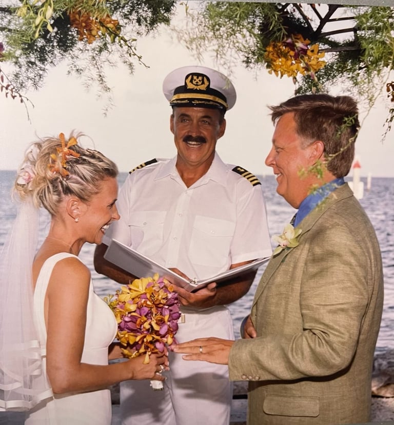 A bride in a white dress with yellow flower lei exchanges vows with a groom in tan jacket while a ship captain in naval uniform officiates the seaside wedding ceremony